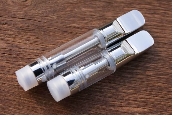 Two Ceramic Cell Vape Cartridges are shown laying lengthwise at an angle against a warm wood textured surface. The cartridges have white mouthpieces and end caps, and a glass body. The longer 1ml cartridge is on top, with the shorter .5ml cartridge below.