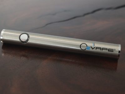 A single 510 Thread battery in stainless steel is laid across a dark rich wood surface. There is a single button on the body, with the O2VAPE logo on the other end.