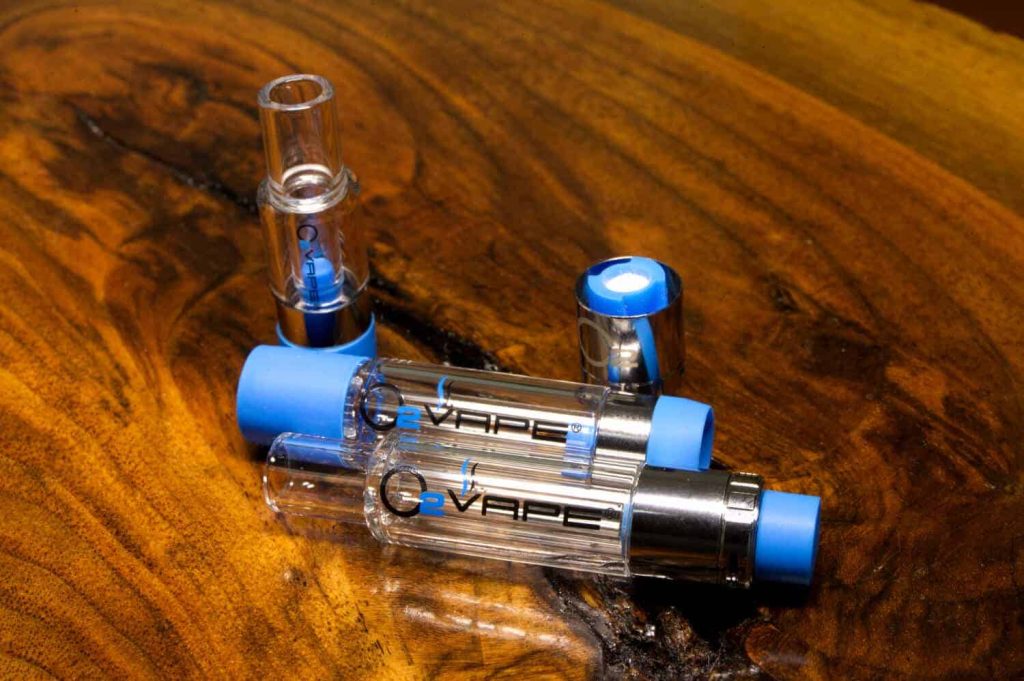 2 All Glass Vari-Flow Vape Cartridges are laid horizontally against a warm wood surface. The cartridge is on top is closed, with blue caps at either end. The cartridge beneath and is open. Above the pens, 2 different sized caps are vertically standing at both ends of the cartridges.