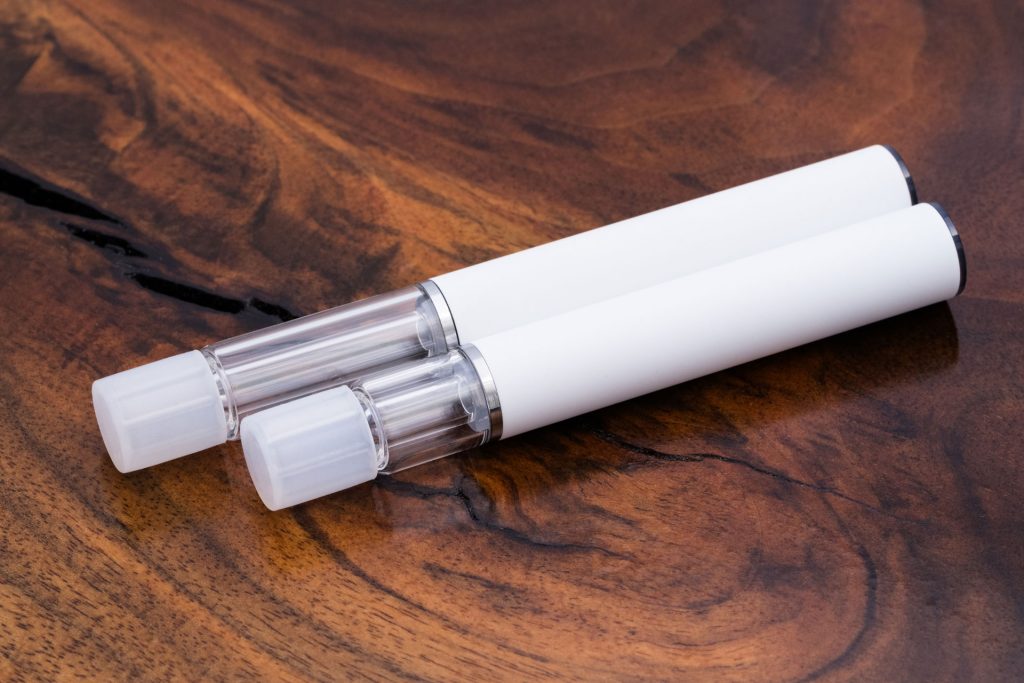 Two Traveler Extreme® All Glass Disposable Vape Pens are resting diagonally on a rich wood surface. The pens are sleek and cylindrical, with a white body over its all-glass construction. The pen in the front is shorter and has a .5ml capacity, while the one in the background is longer and has a 1ml capacity. There are caps inserted into the mouthpieces.