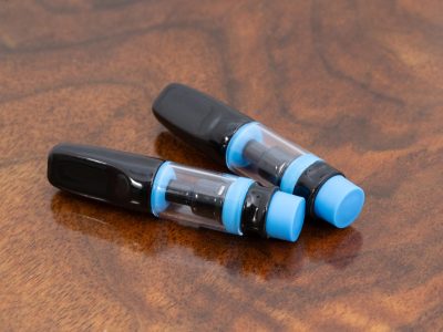 Two blue Full Ceramic vape cartridges with glass bodies are laid horizontally slightly fanned out over a warm wood surface. The mouthpiece caps are black.