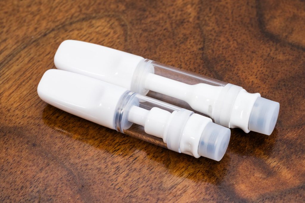 Two white Full Ceramic vape cartridges with glass bodies are laid horizontally at an angle over a warm wood surface. The taller 1ml cartridge is on top, with the shorter .5ml cartridge beneath.