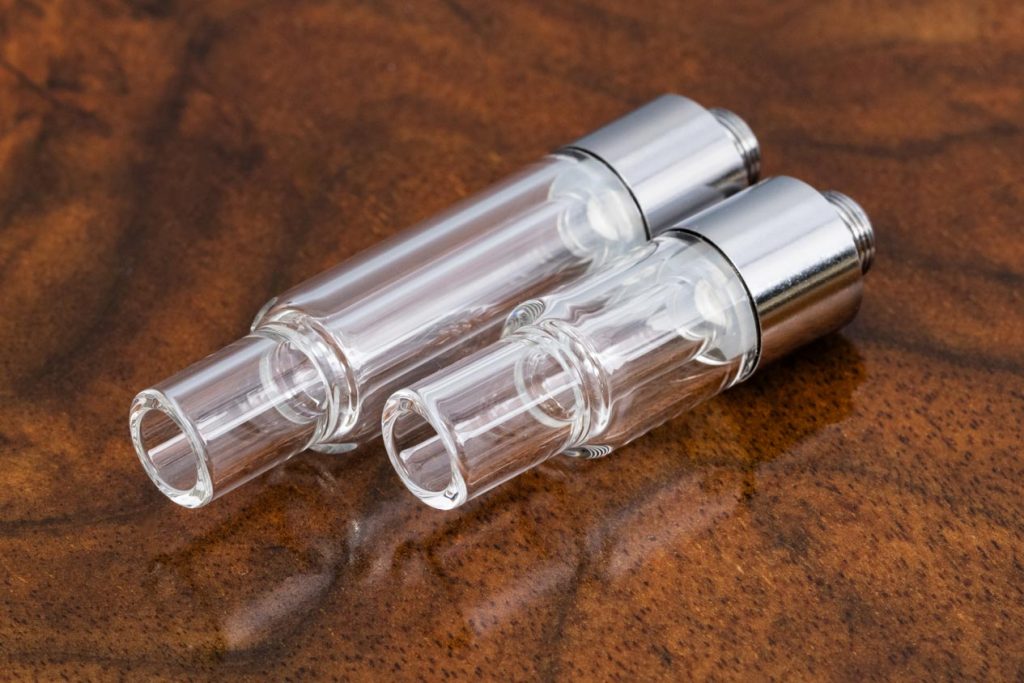 Two All Glass 510 Vape Cartridges are pictured against a warm brown marble surface, displayed at a right angle with the glass mouthpiece at one end, and the base at the other end. The 1ml cartridge on the left is longer, while the smaller .5ml cartridge is on the right.