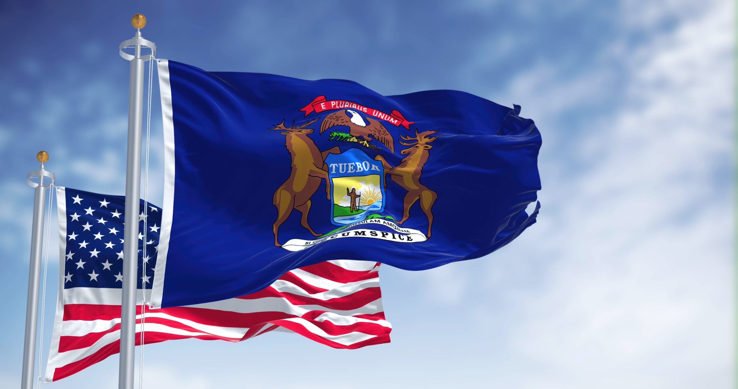 The Michigan state flag waving along with the national flag of the United States of America. In the background there is a clear sky. Michigan is a state in the upper Midwestern of United States