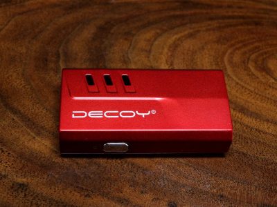 A single red Decoy mini magnetic vape pen lays flat against a dark rich wood surface. The 'DECOY' logo is visible along the front body of the vape pen, with 3 vertical slanted openings above. On the bottom, a micro-USB port is visible.