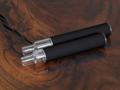 Top-down image of two black unbranded Traveler Extreme All Glass Bucket AIO vape pens with clear glass tanks, shown on a natural wood surface. Wholesale model for discreet, bulk purchase.