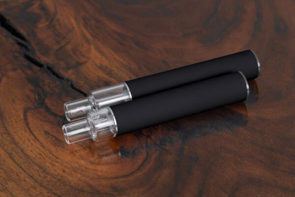 Top-down image of two black unbranded Traveler Extreme All Glass Bucket AIO vape pens with clear glass tanks, shown on a natural wood surface. Wholesale model for discreet, bulk purchase.