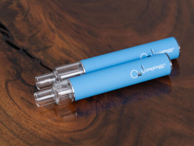 Top-down view of two O2VAPE Traveler Extreme All Glass Bucket AIO vape pens with matte blue finish and transparent all-glass tanks, resting on a natural wood surface. Highlights two different tank sizes in 0.5ml and 1ml.