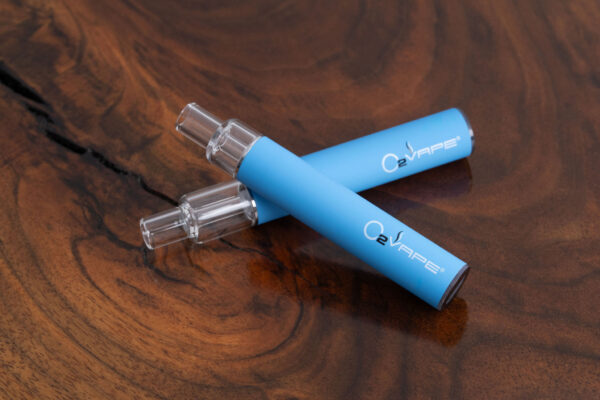 Diagonal layout of two blue O2VAPE Traveler Extreme All Glass Bucket AIO devices crossed on a rich wood surface, emphasizing the minimalist all-glass construction.