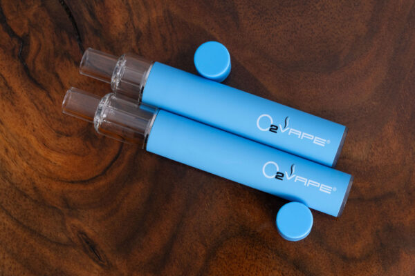 Angled top down view of two O2VAPE Traveler Extreme All Glass Bucket AIO vape pens stacked with visible clear tanks, center posts, and sleek matte blue bodies. Clean, discreet, and resin-ready design showcased.