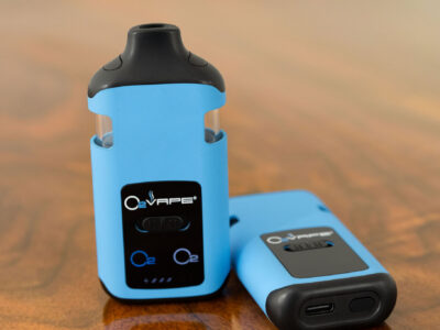 Two O2VAPE Traveler Extreme Glass Dual vape devices in blue, one standing upright showing front display with dual O2 chamber indicators and all-glass tank, one laying flat showing USB-C charging port and power button
